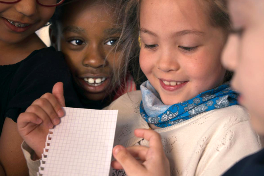 4 enfants souriants, les uns contre les autres, regardent une feuille à petits carreaux. 