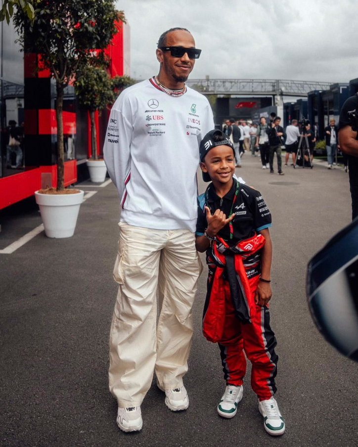 Amine, à côté de Lewis Hamilton. Lewis Hamilton porte un sweat blanc et un pantalon couleur crème. Amine, petit à côté de lui, est en combinaison rouge, avec une casquette noire sur la tête. Il est très fier, et fait le signe des cornes avec sa main droite. C'est un signe de fan, de complicité.