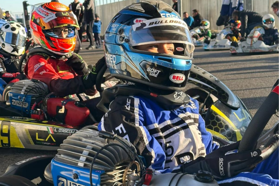 Au premier plan, Amine assis dans son kart. Il porte une combinaison bleue et un gros casque bleu sur la tête. Il est concentré. Derrière lui, d'autres enfants dans leur kart.